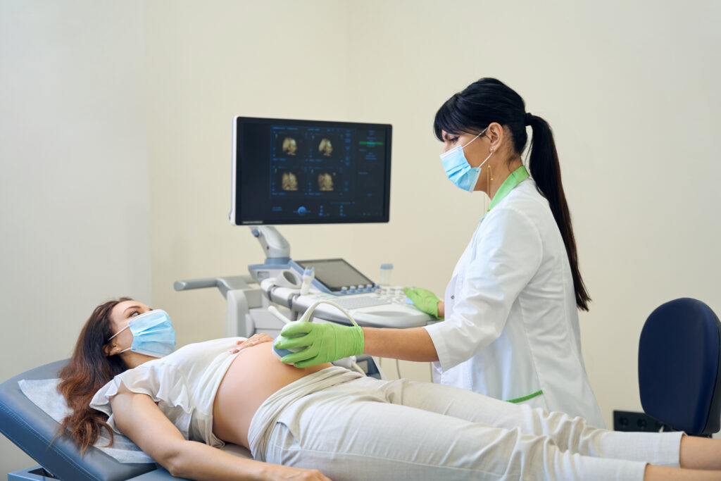 Adult european female gynecologist doing ultrasound examination of young caucasian pregnant woman in clinic. Females wearing medical masks. Concept of pregnancy examination